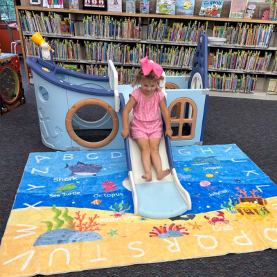 photo with library shelves in the backgroun and a girl sliding down a toy boat onto a beach-themed carpet with letters on it