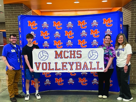Nick Adams of the library stands in front of a MC backdrop with two students and a teacher. The students are holding a vinyl MCHS Volleyball sign made in the library's MakerSpace.