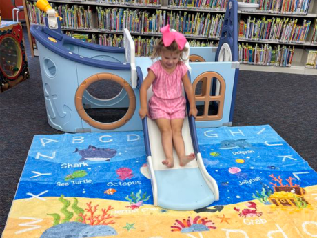 photo with library shelves in the backgroun and a girl sliding down a toy boat onto a beach-themed carpet with letters on it