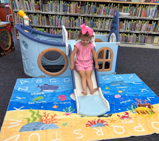 photo with library shelves in the backgroun and a girl sliding down a toy boat onto a beach-themed carpet with letters on it