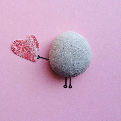 a round rock on a pink piece of paper. legs and an arm have been drawn onto the paper to make the rock look like a person, and he has a piece of red paper cut into the shape of a heart in his hand